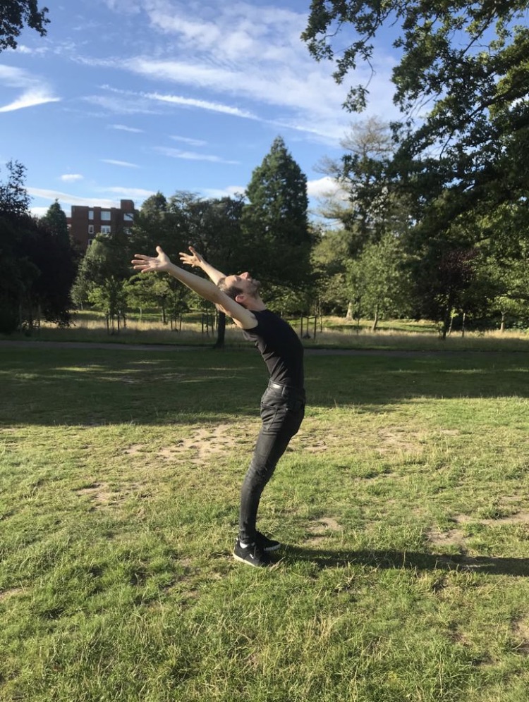 Angelo in a park performing a stretch with arms extended towards the sky.